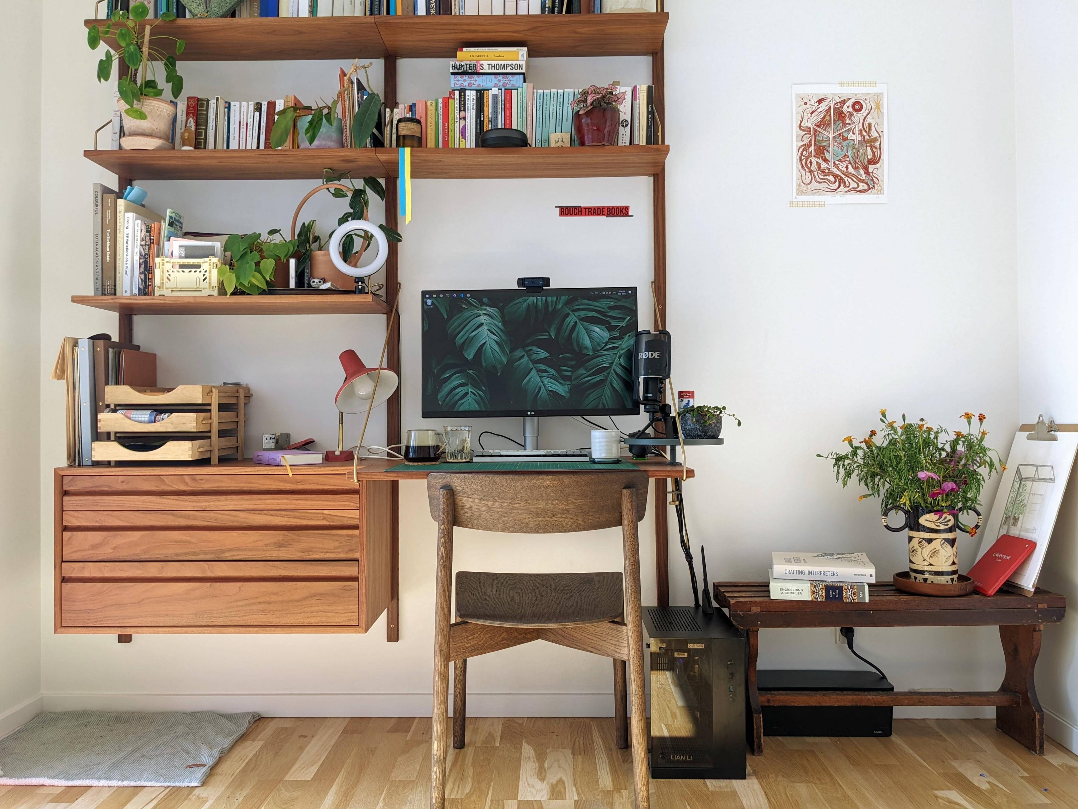 The photo depicts a walnut bookshelf system against a white wall. Part of this system is a desk with a large monitor showing a backdrop of vivid monstera leaves. The shelf is full of various colourful books, nicknacks, and plants. Right of the book shelf standing on the floor is a small black box, a self-built computer. Next to is a vintage-looking bench with several more books, and black vase with flowers. There is a bit of sunshine coming in from the left of the photo, giving this space a welcoming and warm vibe.